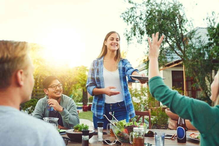 Group of friends enjoying a sunny outdoor meal in a garden setting, woman serving food and smiling warmly